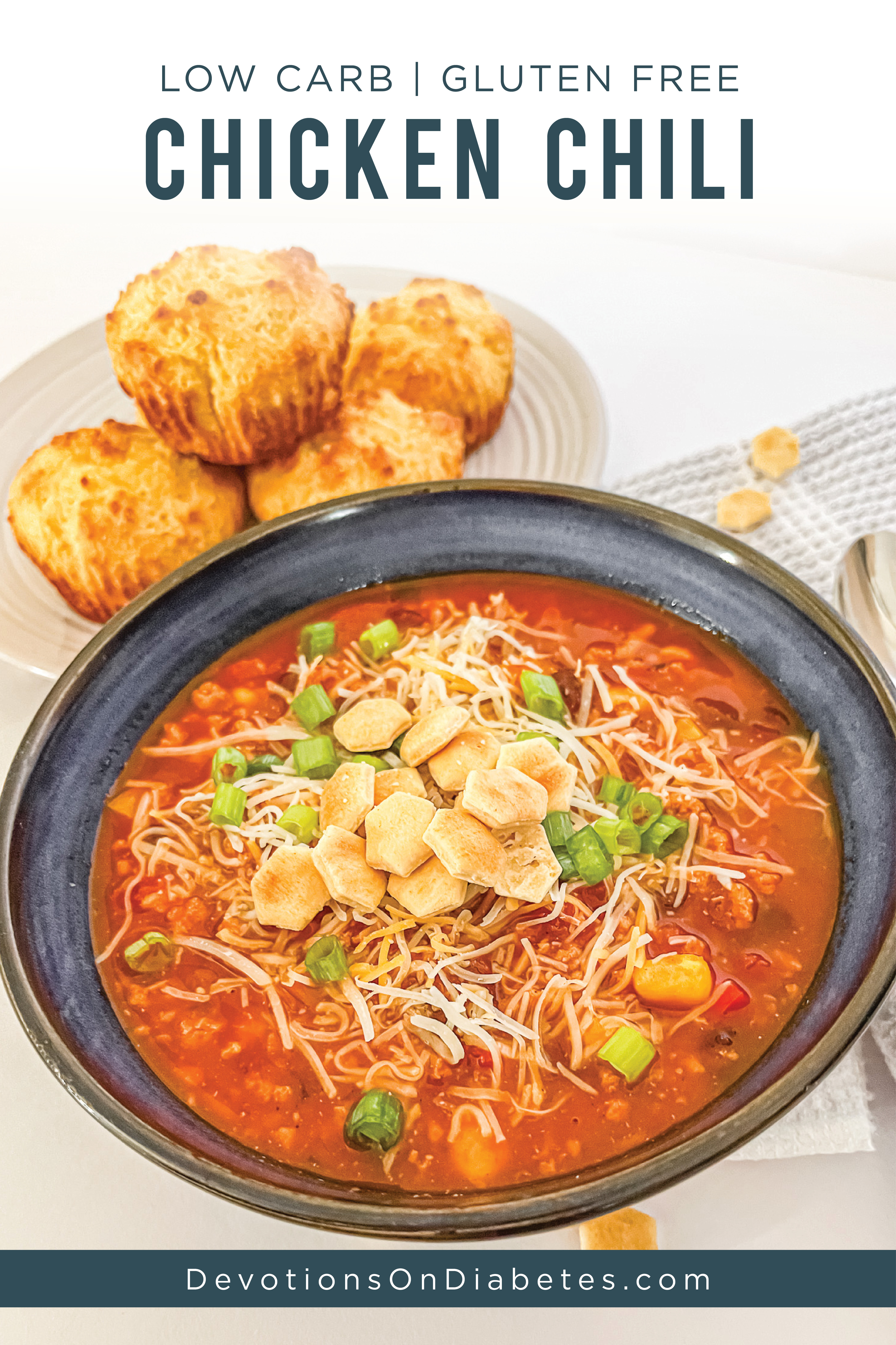 Low Carb Gluten Free Chicken Chili. Pictured: a bowl of chili with cheese and green onion and crackers with cornbread muffins on the side.